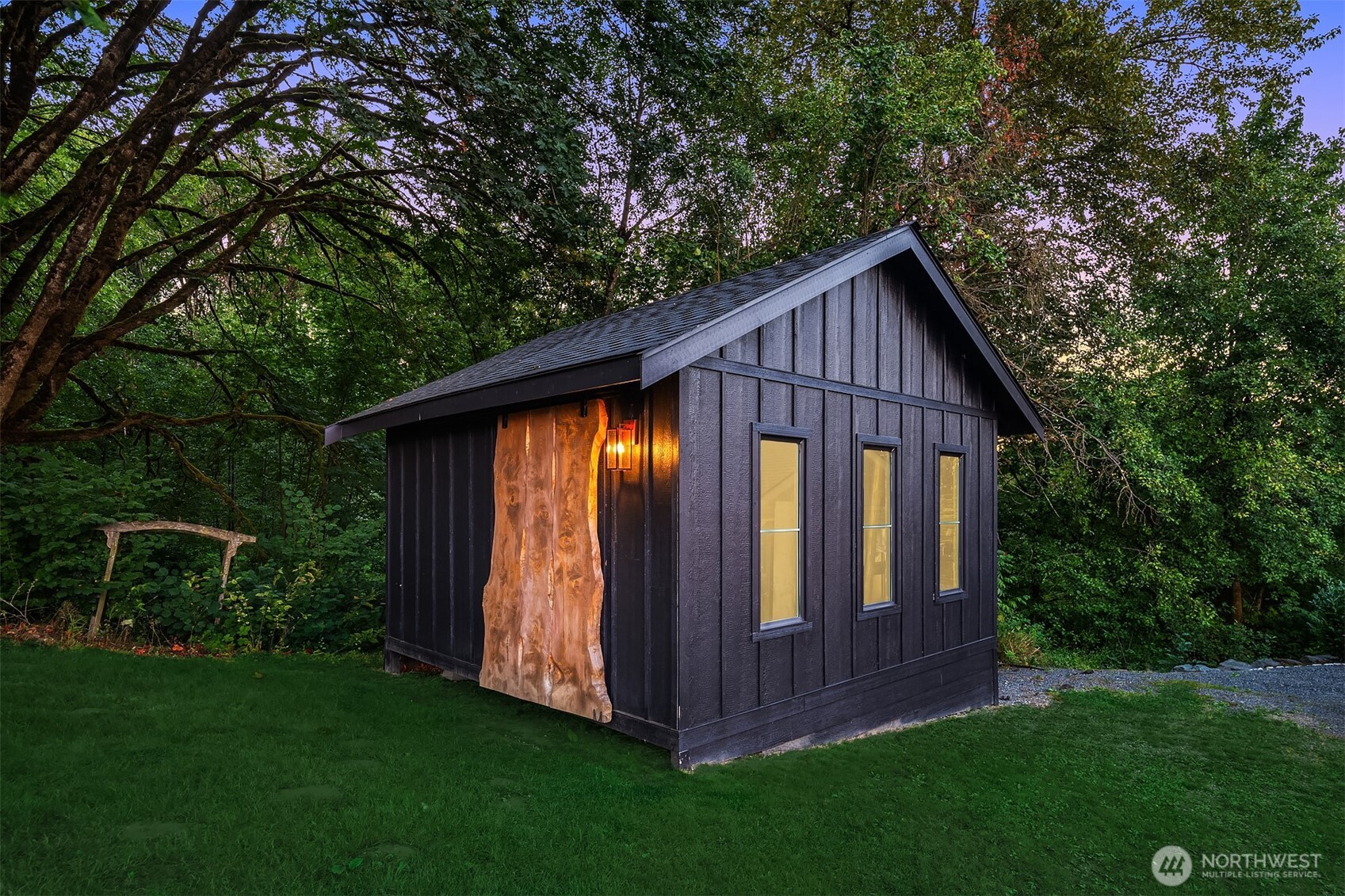 Black outbuilding with a live-edge wooden door and glowing lantern beneath mature trees.