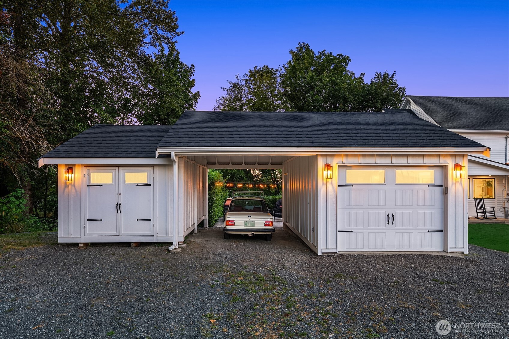 White detached garage with a covered drive-through carport and a car parked beneath it at dusk.