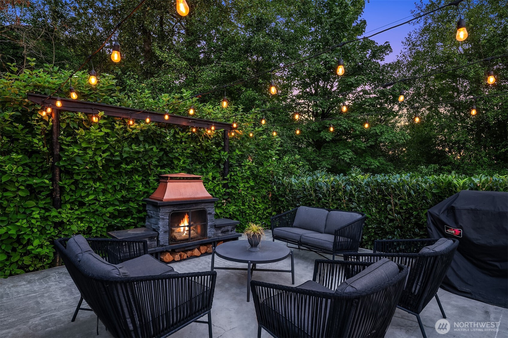 Outdoor patio with cushioned seating, a lit stone fireplace, and string lights among leafy hedges.