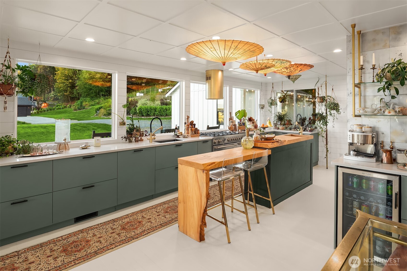 Staged kitchen with dark green cabinets, a large wood island, hanging parasols, and garden views through wide windows.