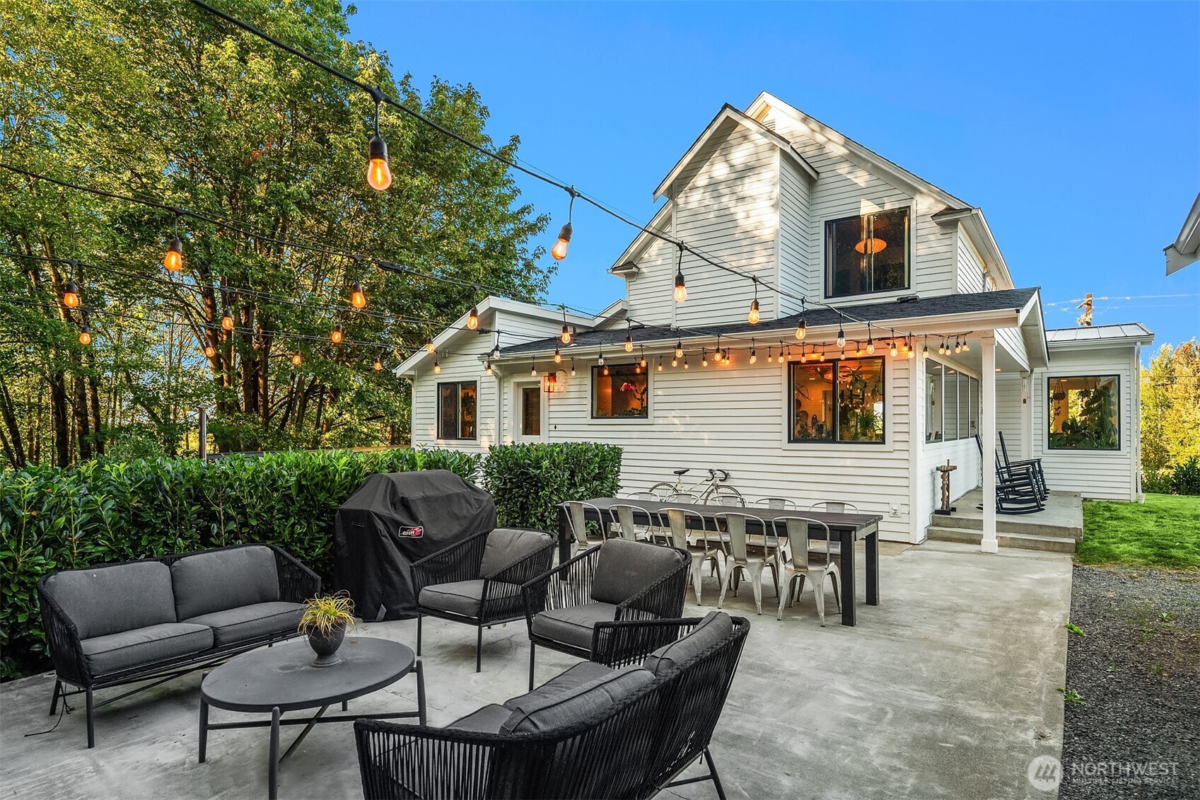 White-sided home with a furnished concrete patio, outdoor dining table, and overhead string lights.