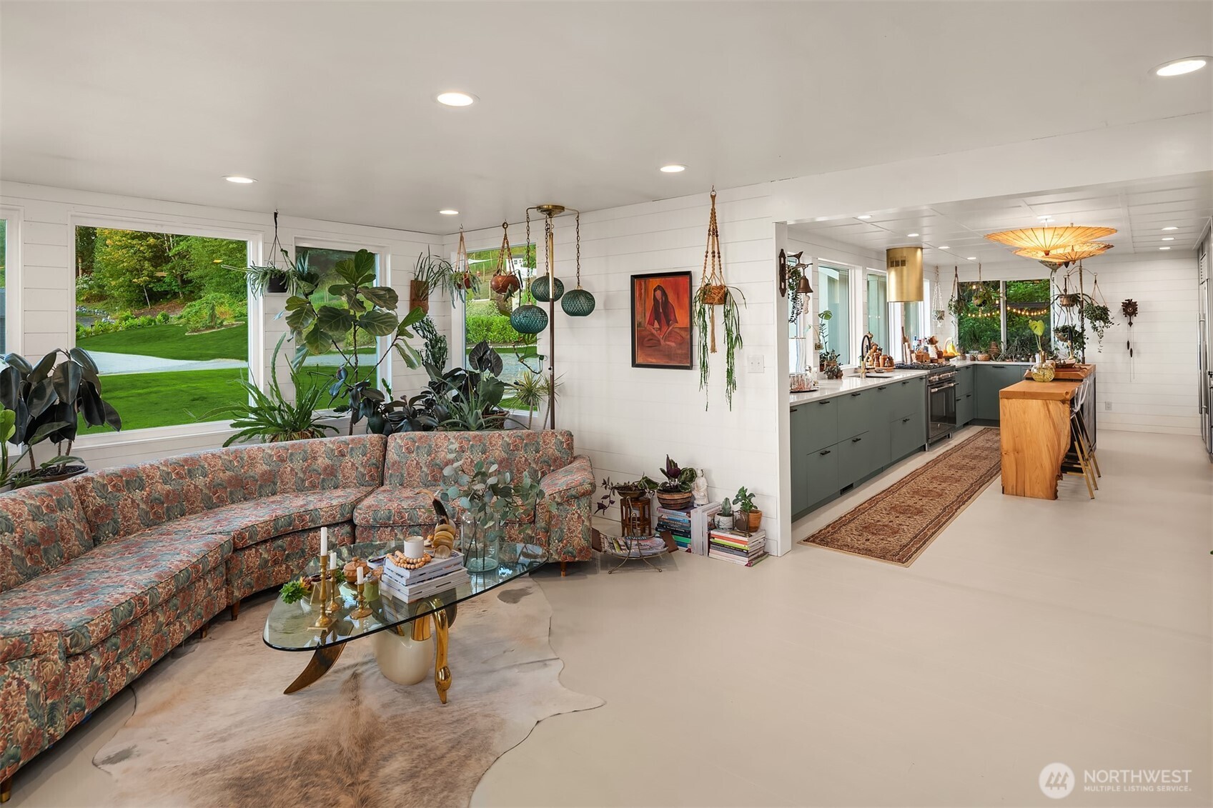 Open-plan living room and kitchen with a floral sectional, abundant plants, and green cabinetry.