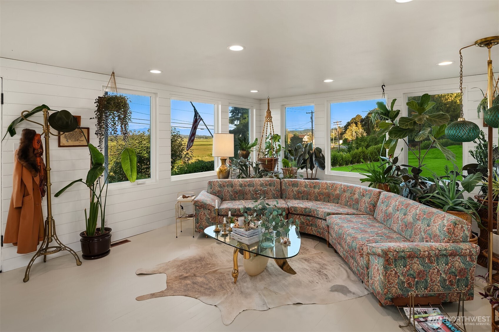 Living room with a floral sectional, abundant plants, and large windows overlooking lawns and fields.