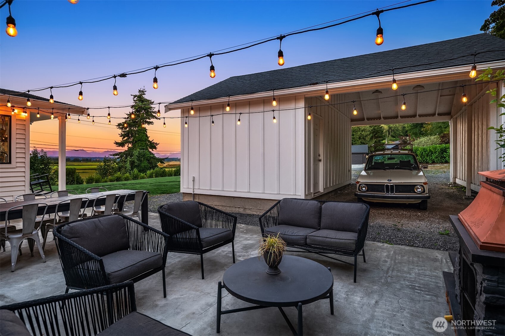 Patio with string lights, lounge seating, covered carport, and distant fields at dusk.