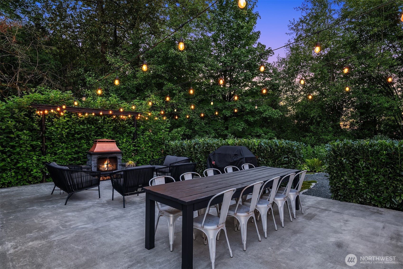 Furnished patio with a long dining table, glowing string lights, and a lit stone fireplace among tall hedges.