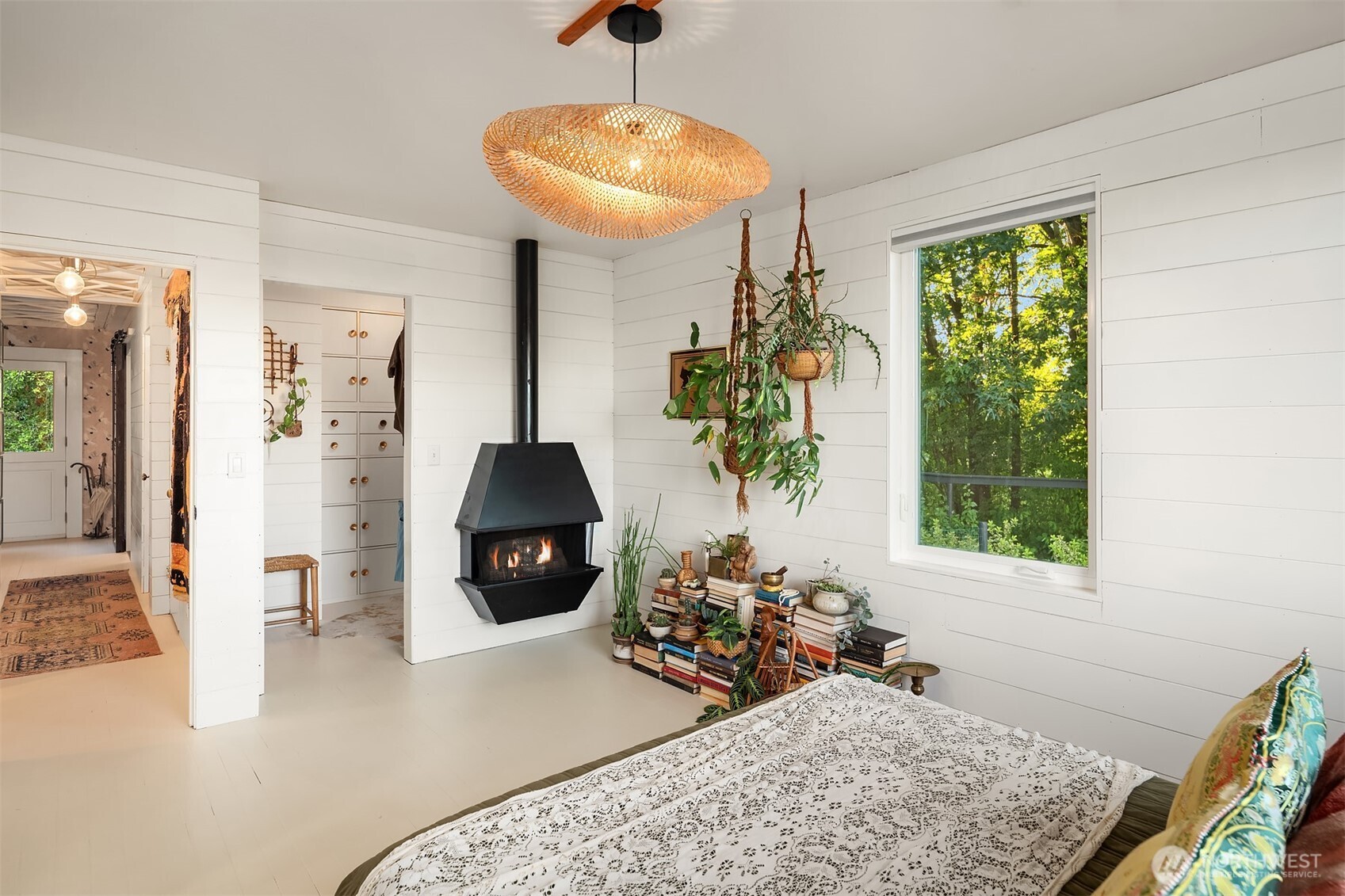 Bedroom with a black freestanding fireplace, woven pendant light, and large window overlooking trees.