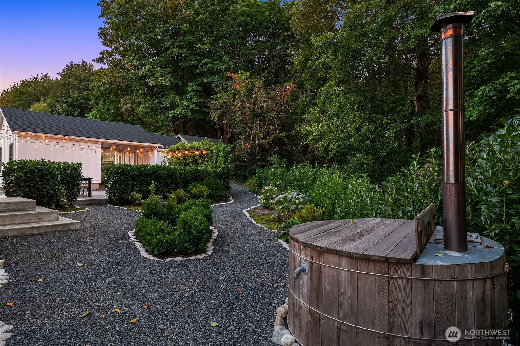 Wood-fired soaking tub beside gravel garden paths and a light-strung white outbuilding.