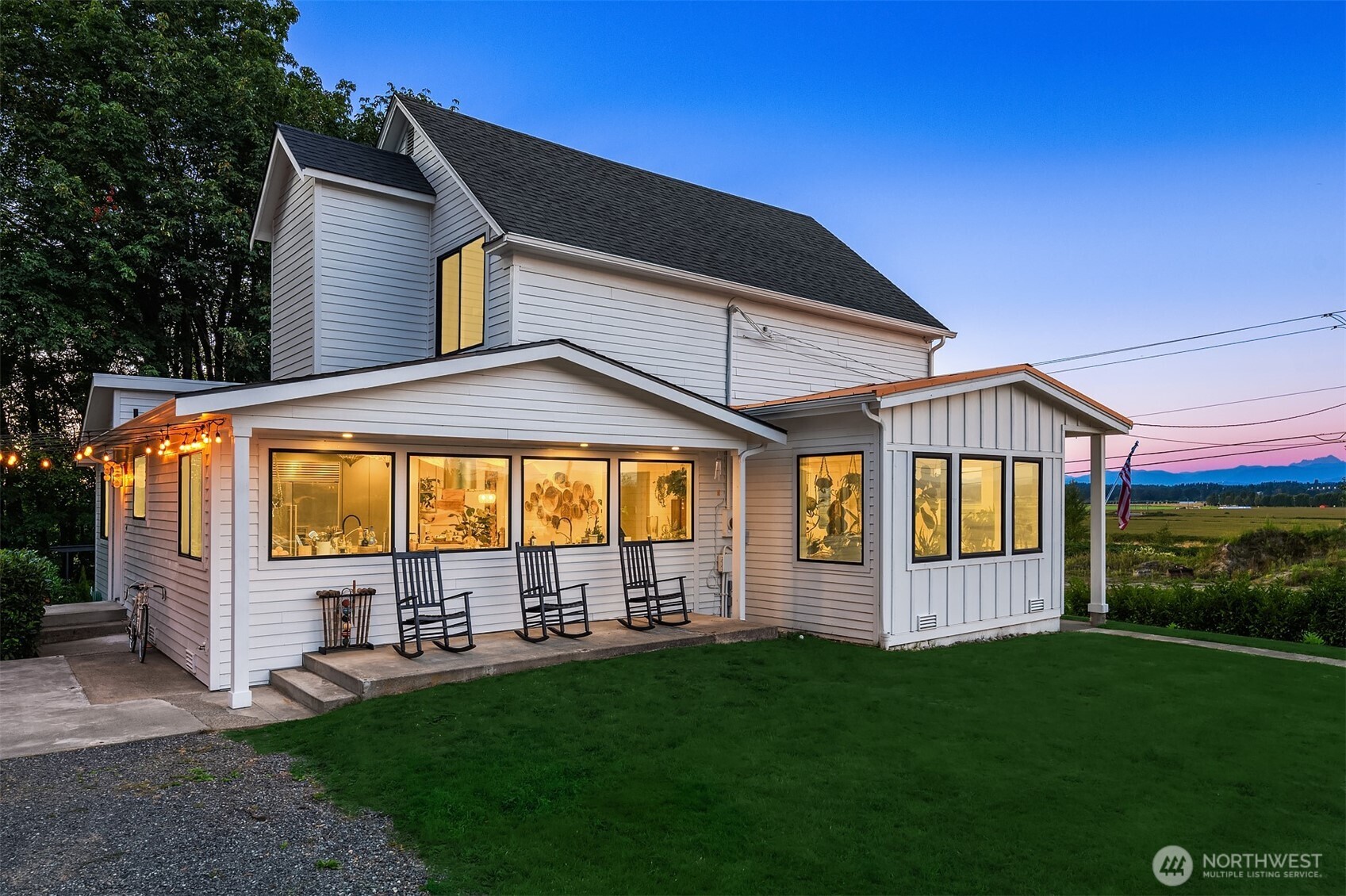 White two-story home with a covered porch, rocking chairs, warm windows, and distant mountain views at dusk.