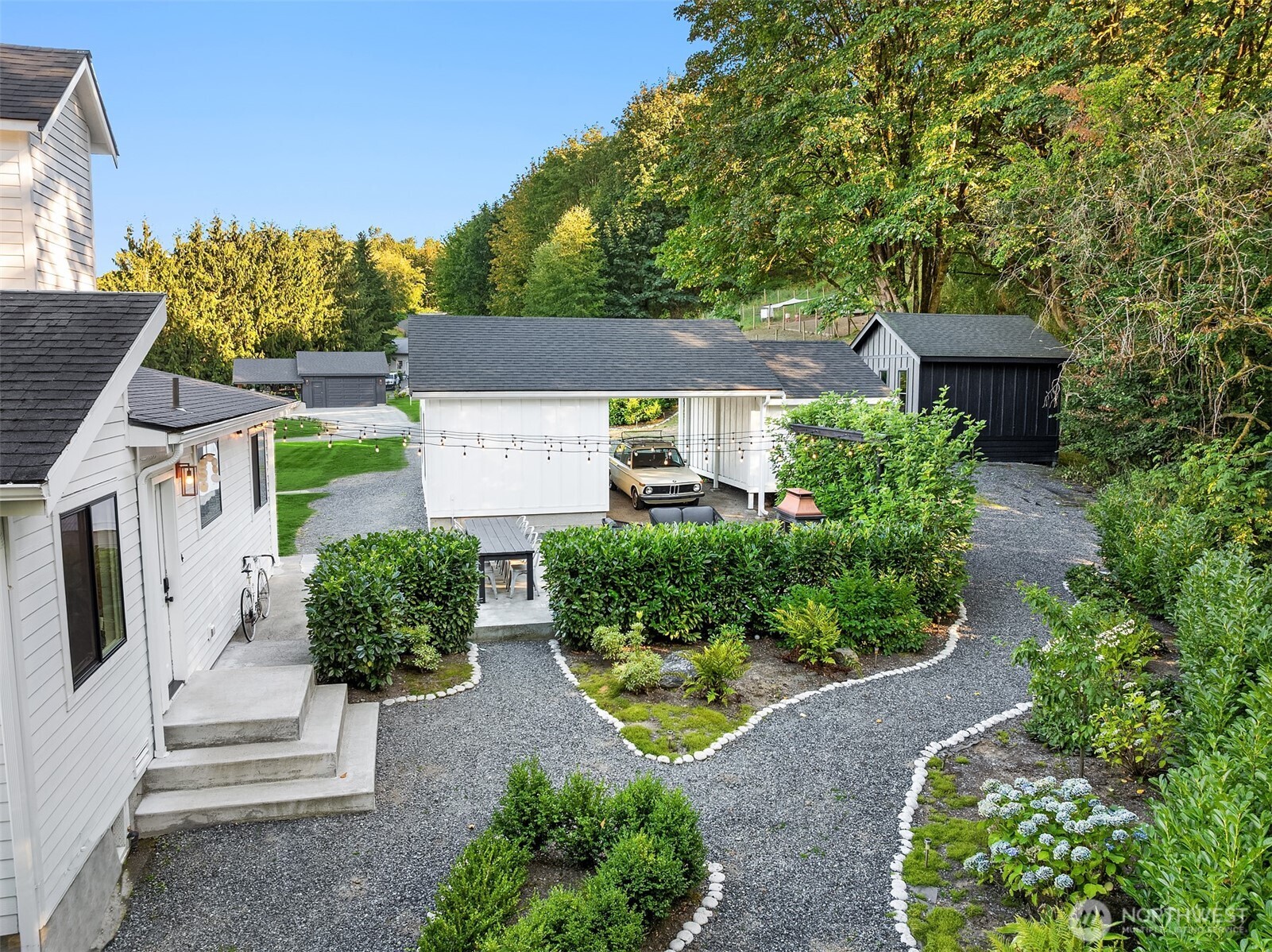 Residential courtyard with gravel paths, trimmed hedges, white outbuildings, and a vintage car.