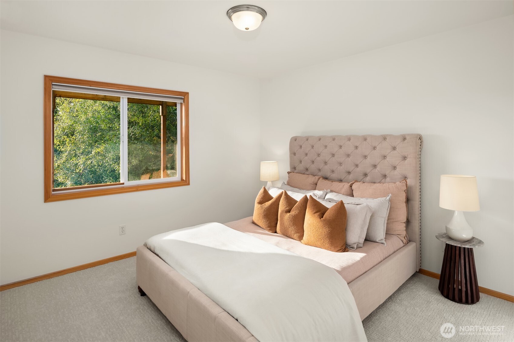 Staged bedroom with a blush-pink tufted bed, rust-colored pillows, and a wood-trimmed window overlooking greenery.