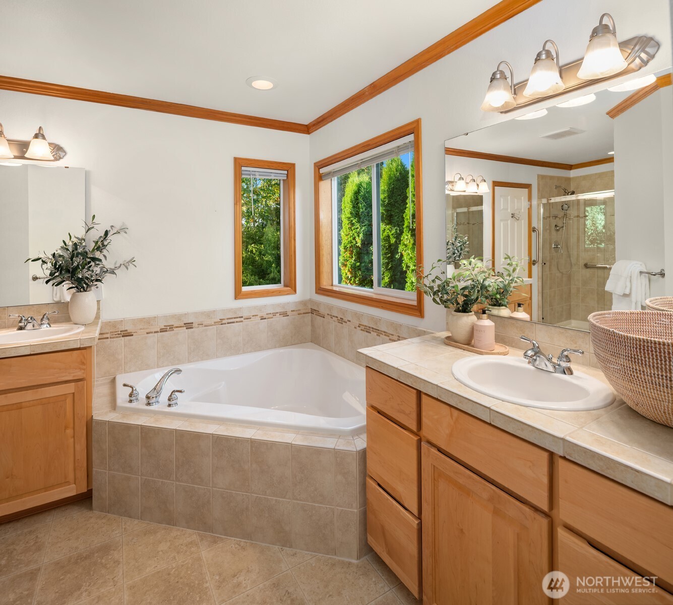 Bathroom with a tiled soaking tub, wood cabinetry, and windows overlooking greenery.