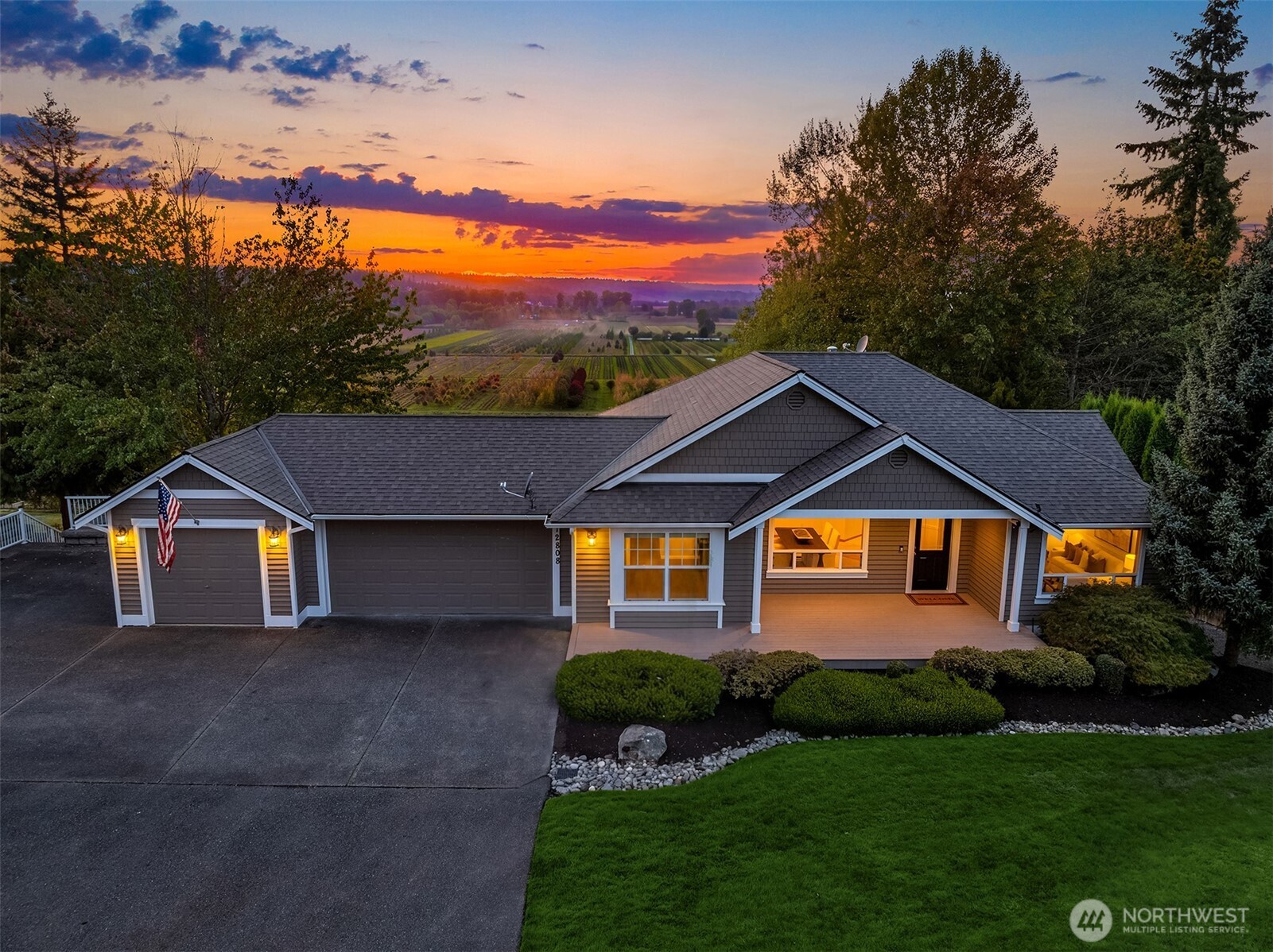 Gray single-story home with a three-car garage and covered porch at sunset, overlooking farmland.