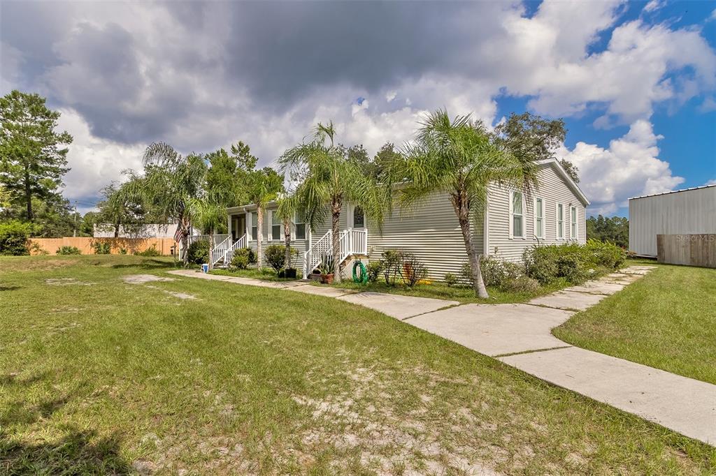 Single-story light-sided home with a covered porch, palm trees, grassy yard, and concrete driveway.