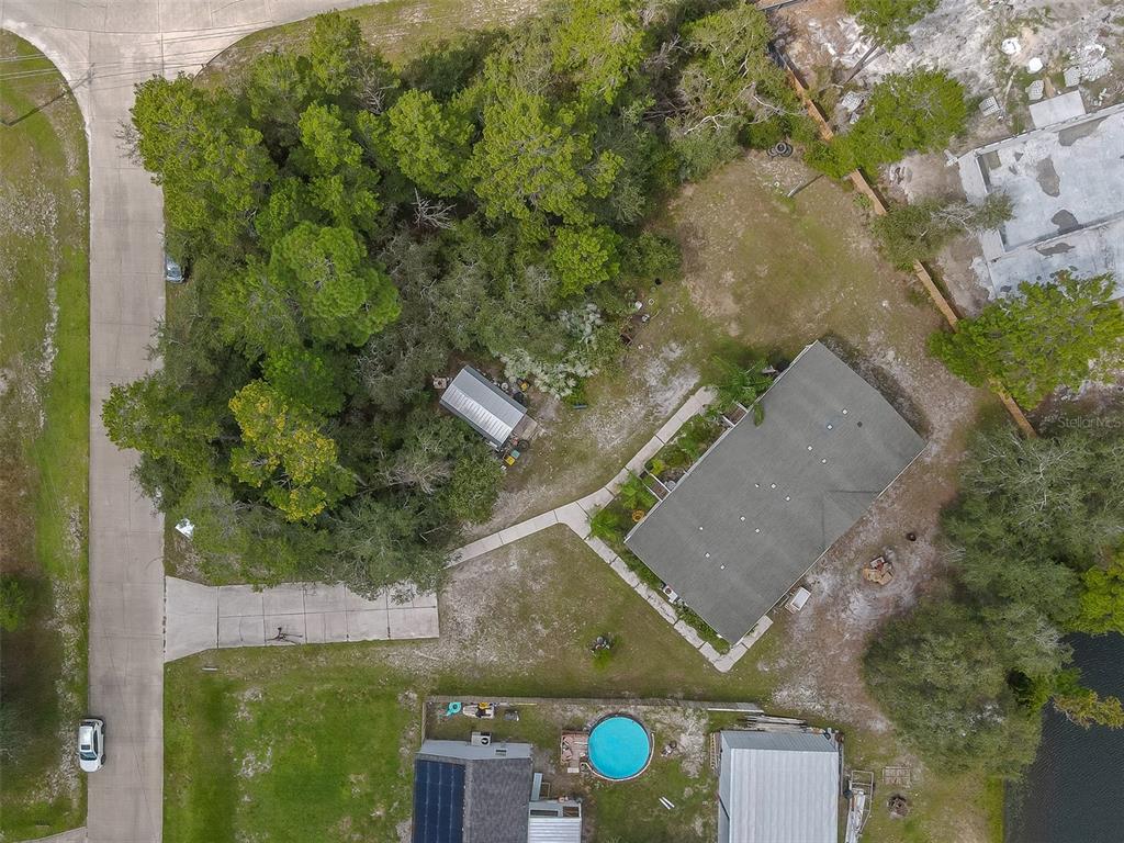 Aerial view of a wooded property with a gray-roofed house, blue pool, driveway, and shoreline.