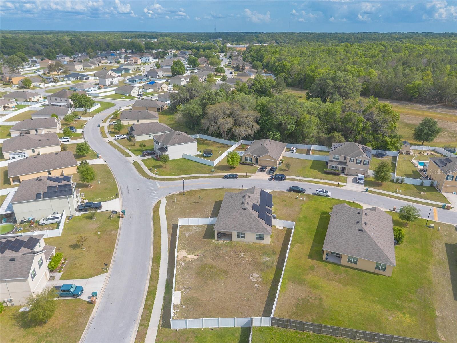 Aerial view of a fenced vacant lot among suburban homes, with woods behind the neighborhood.