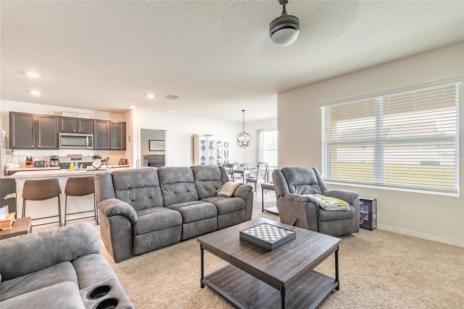 Open-plan living room with gray reclining sofas, carpeted floors, and a kitchen and dining area beyond.