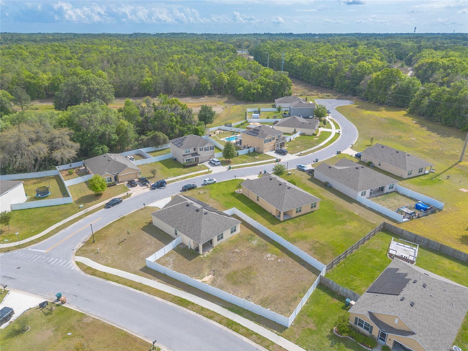 Aerial view of a fenced vacant lot among neighborhood homes, with woods beyond.