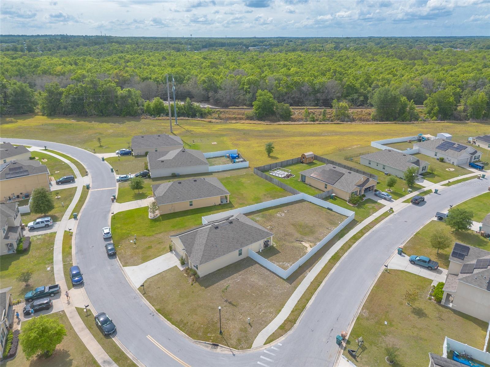 Aerial view of a fenced vacant lot among homes, with a wooded area beyond the neighborhood.