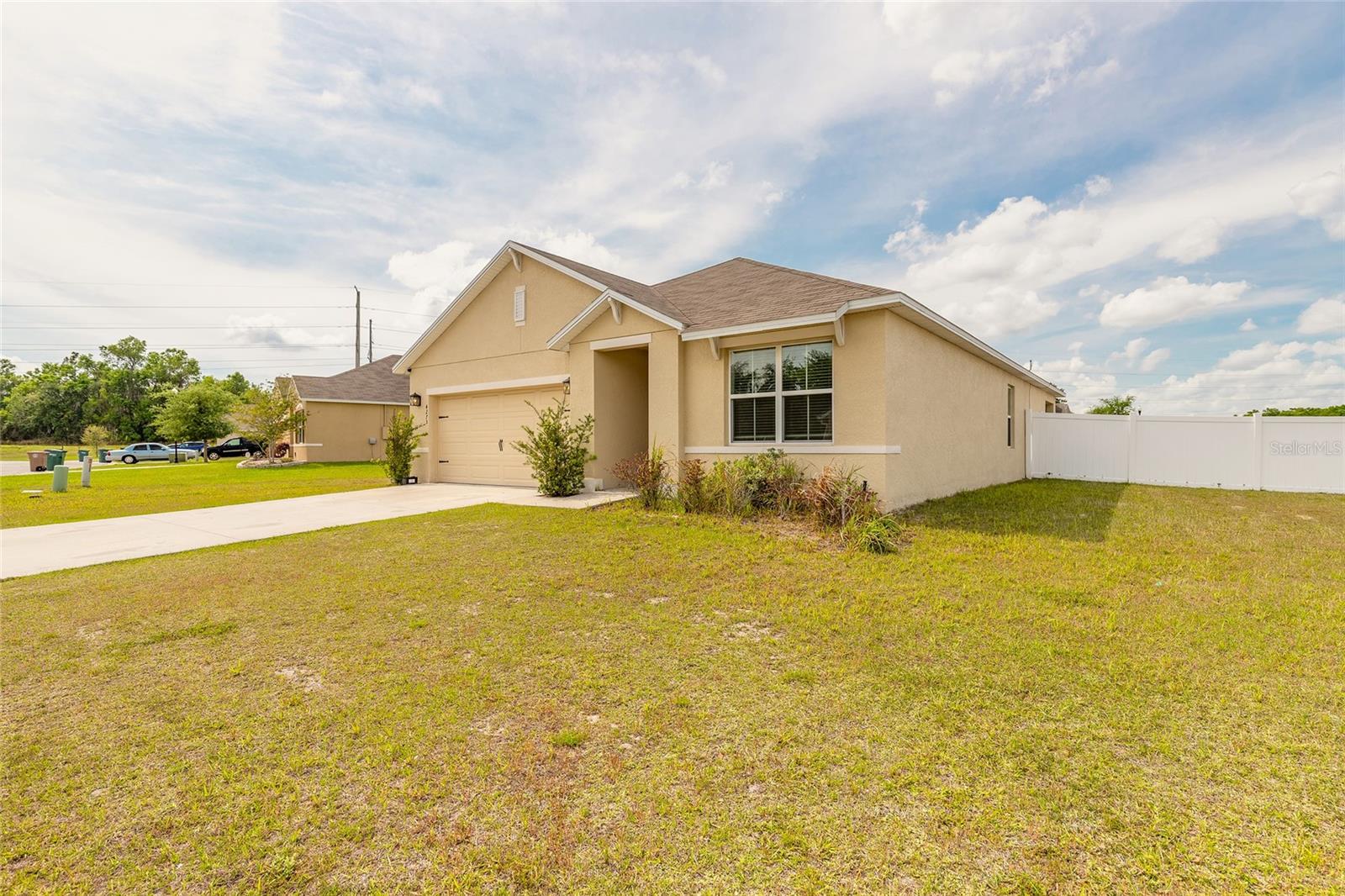 Beige single-story home with a two-car garage, broad driveway, lawn, and white side-yard fence.