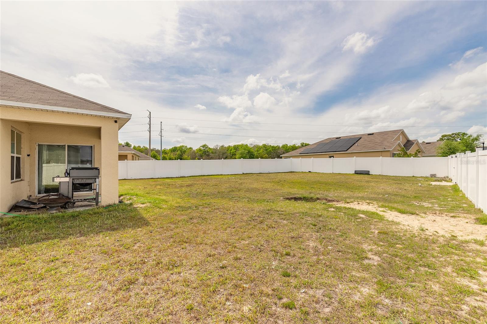 Fenced backyard with patchy grass, a barbecue grill, and neighboring homes with rooftop solar panels.