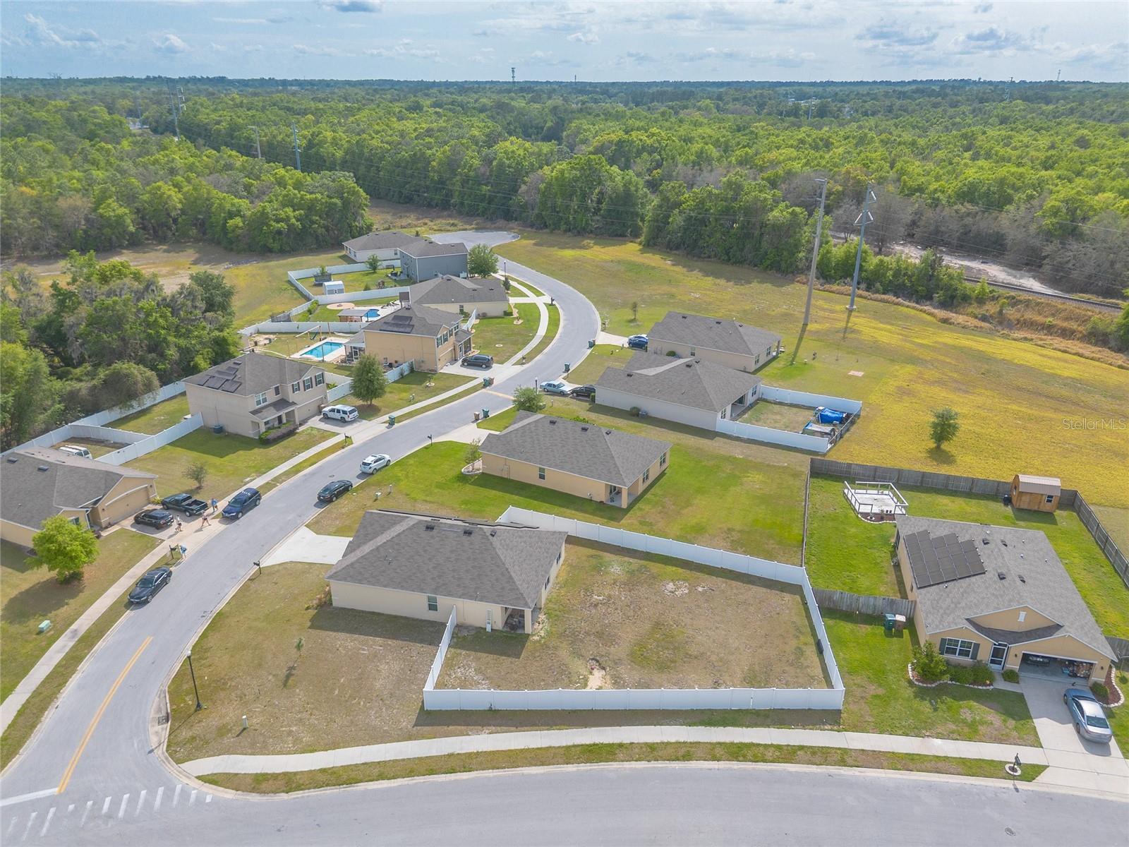 Vacant fenced lot beside a curved neighborhood street, with homes and woods nearby.