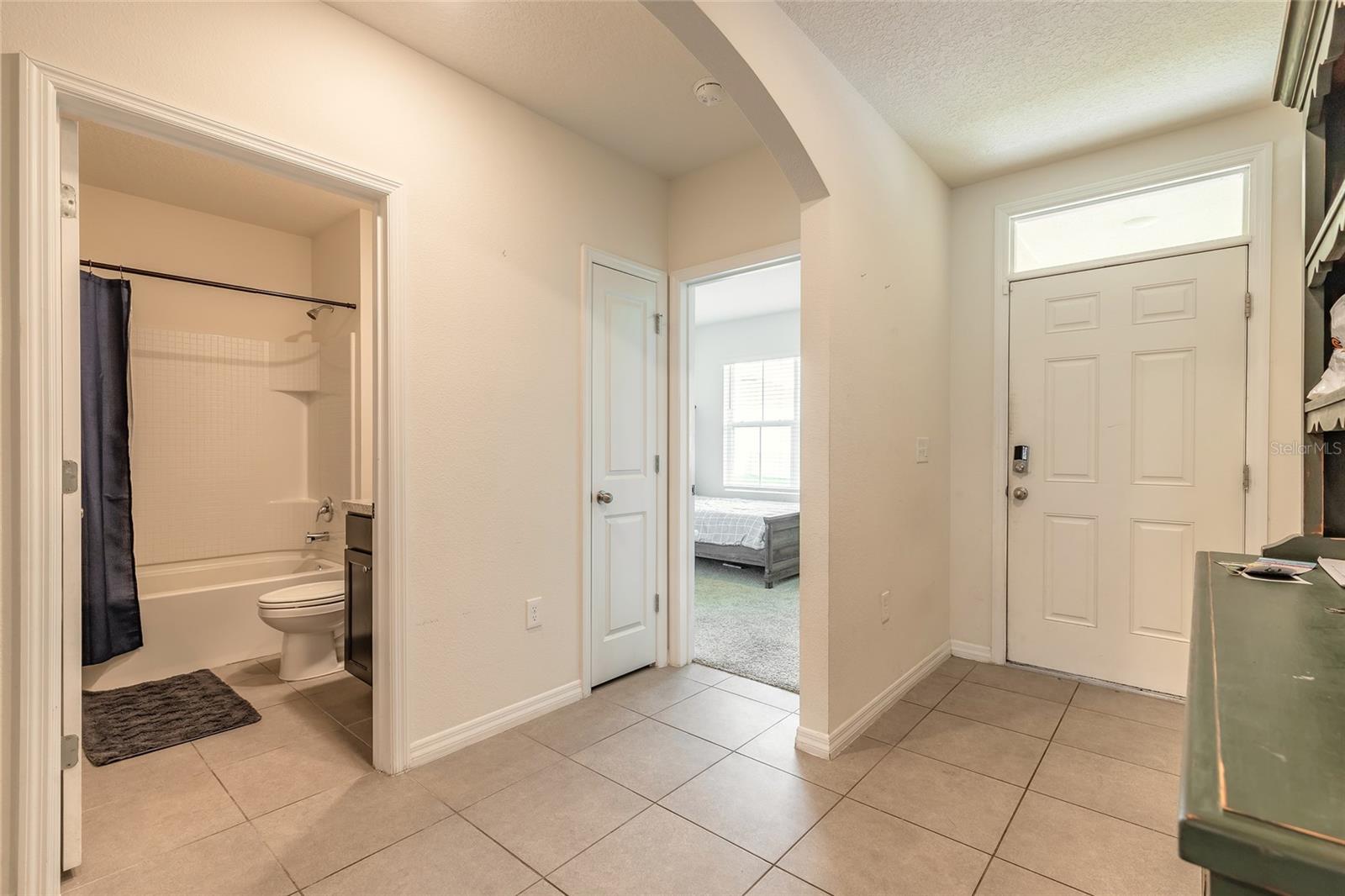 Tiled entry hall with the front door, an open bathroom, and a bedroom visible through a doorway.
