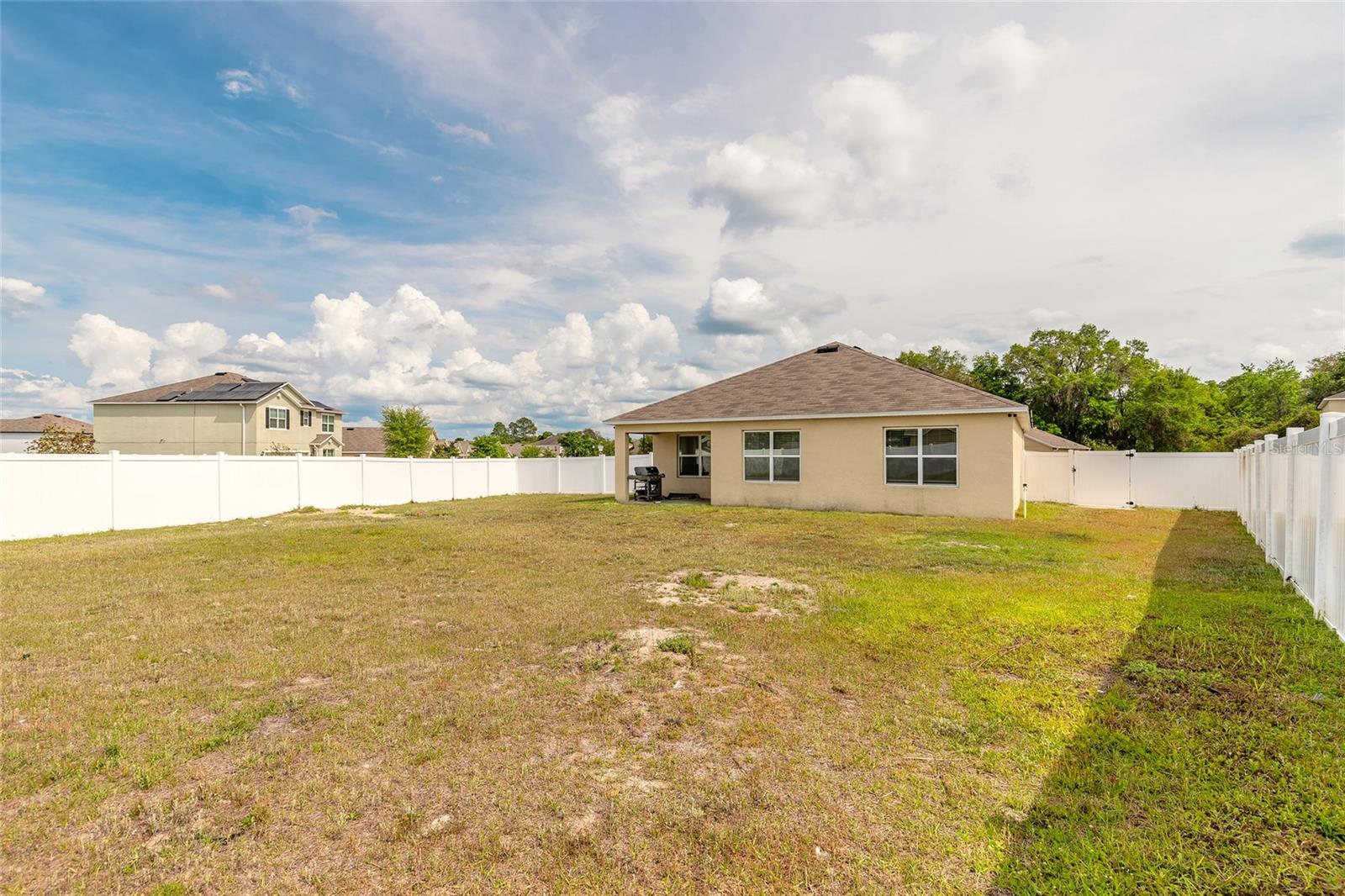 Fenced backyard with a wide grassy lawn, white vinyl fence, and the rear of a beige house.