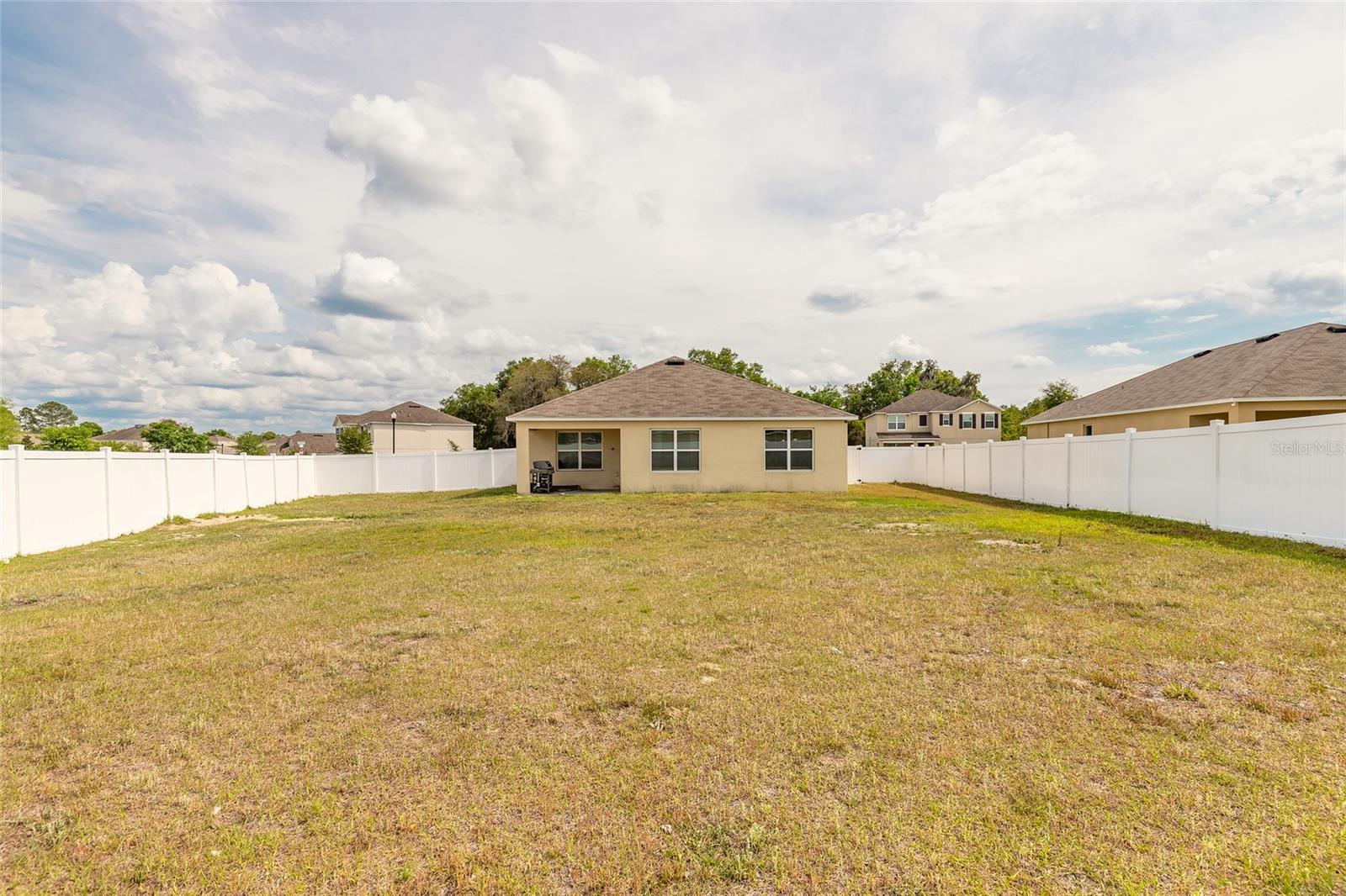 Backyard with a broad lawn enclosed by white vinyl fencing behind a single-story beige home.