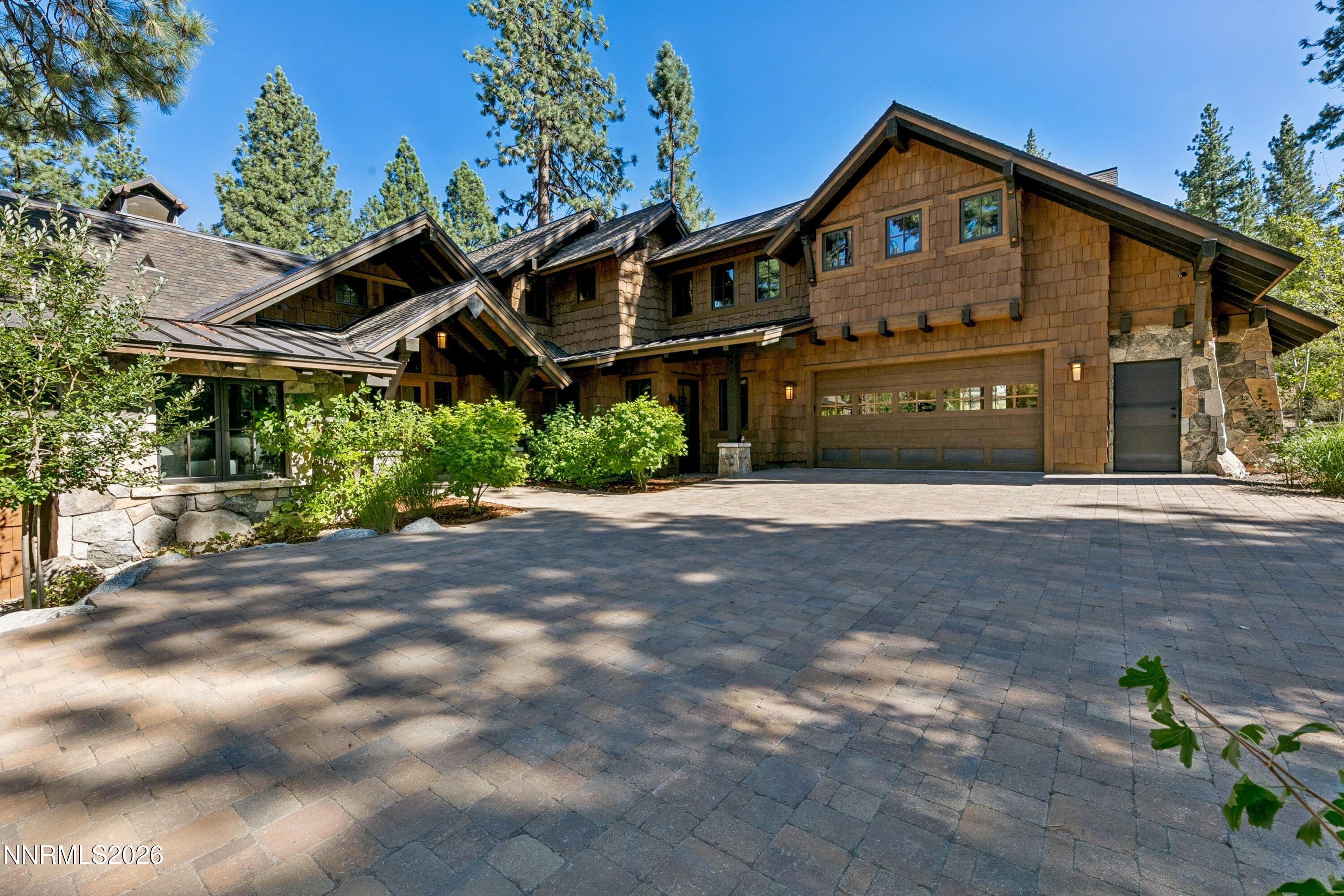 Two-story shingle home with stone accents, a wide paved driveway, and tall pines.