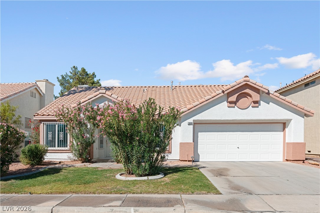 Single-story stucco home with a tiled roof, flowering shrubs, front lawn, and attached garage.