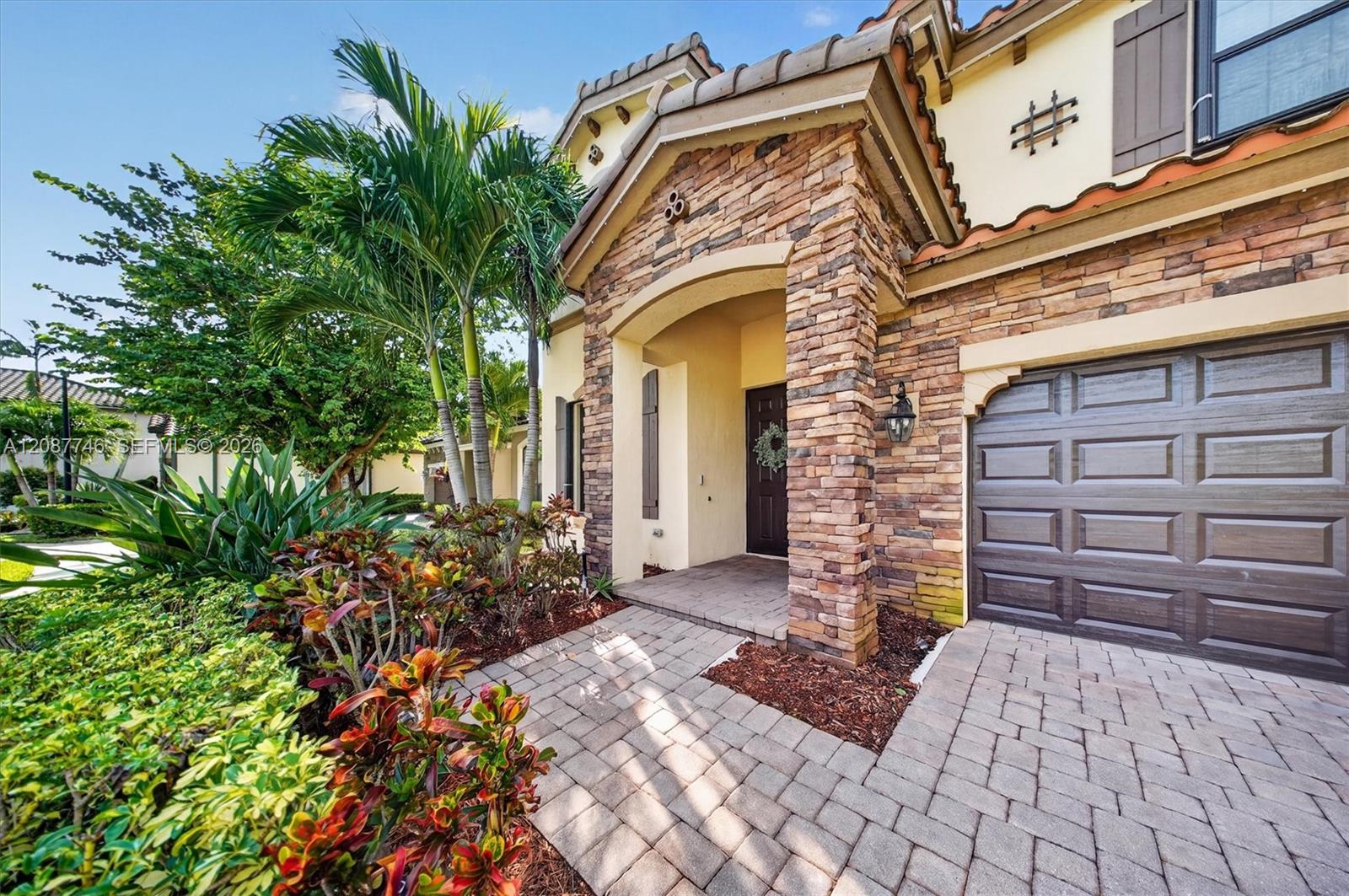 Home exterior with a stone archway, dark garage door, tropical landscaping, and paver driveway.