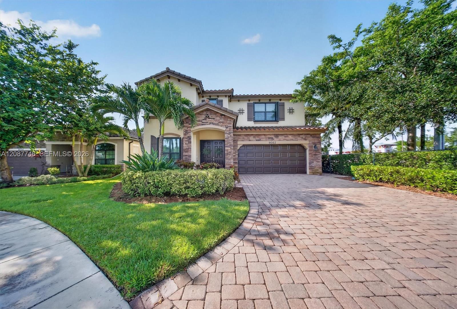 Two-story home with stone-accented entry, palm trees, and a wide paver driveway.