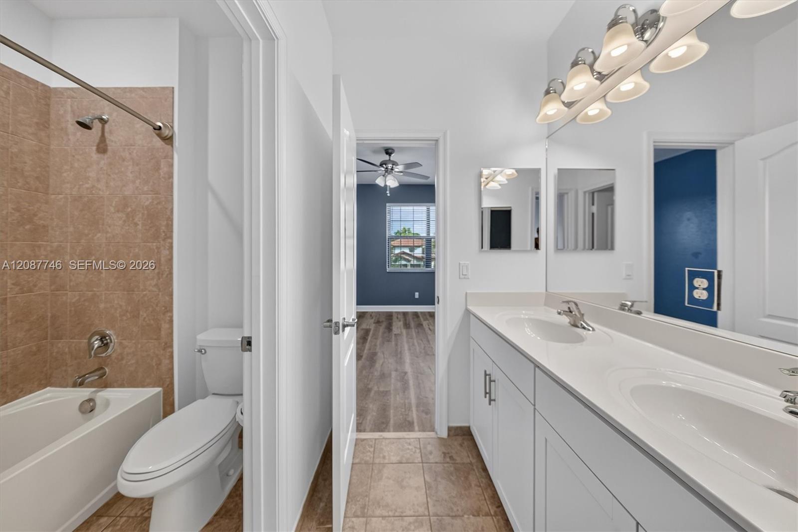 Bathroom with a white double vanity, tan-tiled bathtub surround, and open doorway to a blue room.