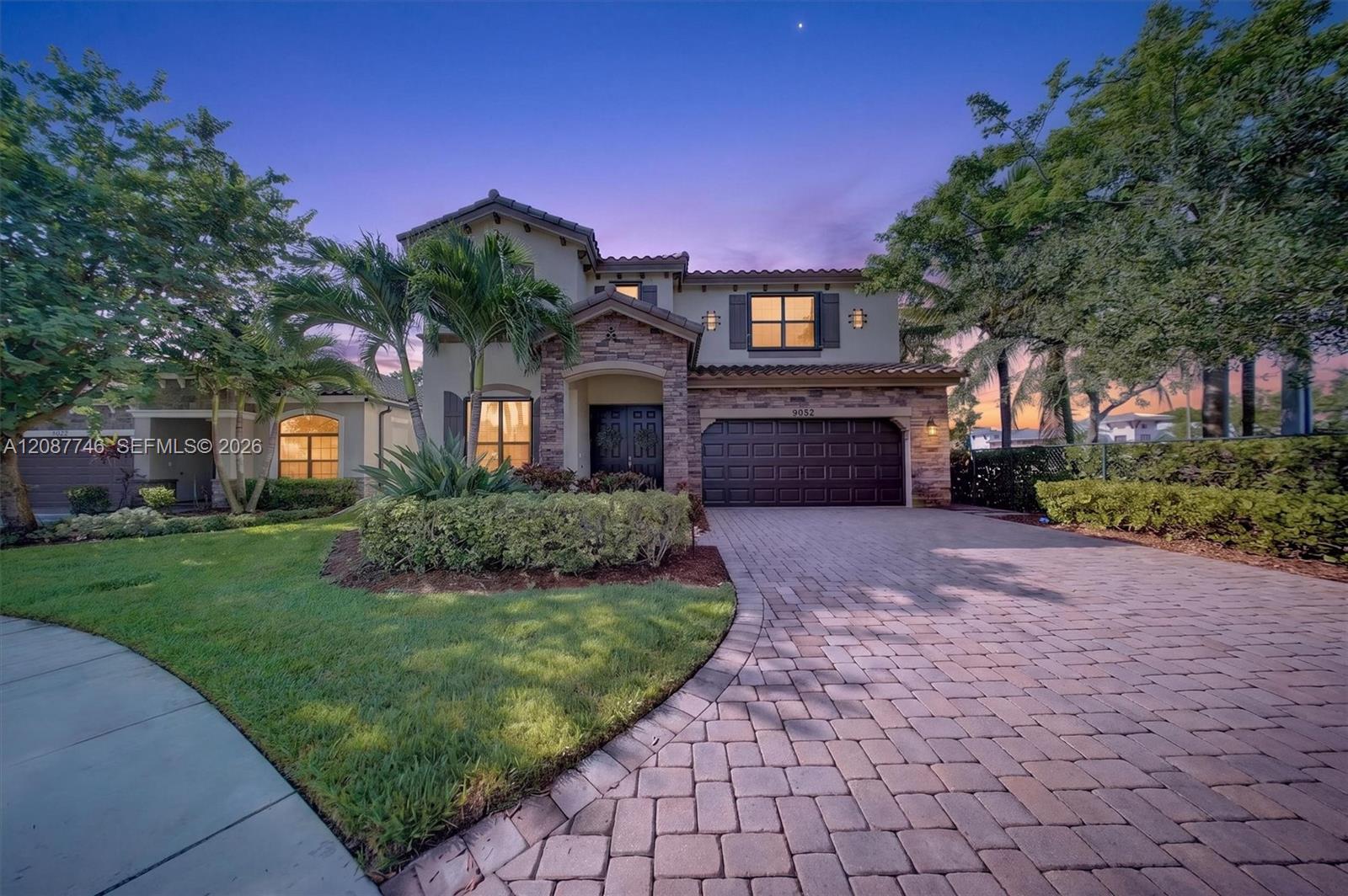 Two-story home with stone accents, palm landscaping, and a wide paver driveway at dusk.