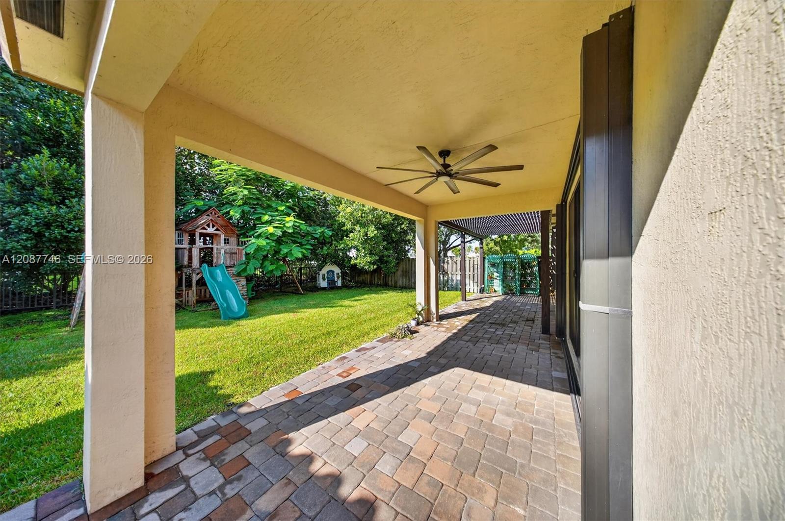 Covered paver patio with ceiling fan overlooking a fenced backyard and children's playset.