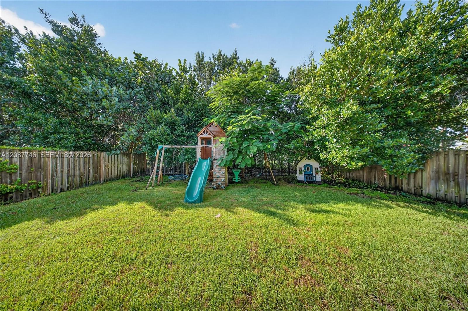 Fenced backyard with a wooden playset, teal slide, swings, and a small playhouse.