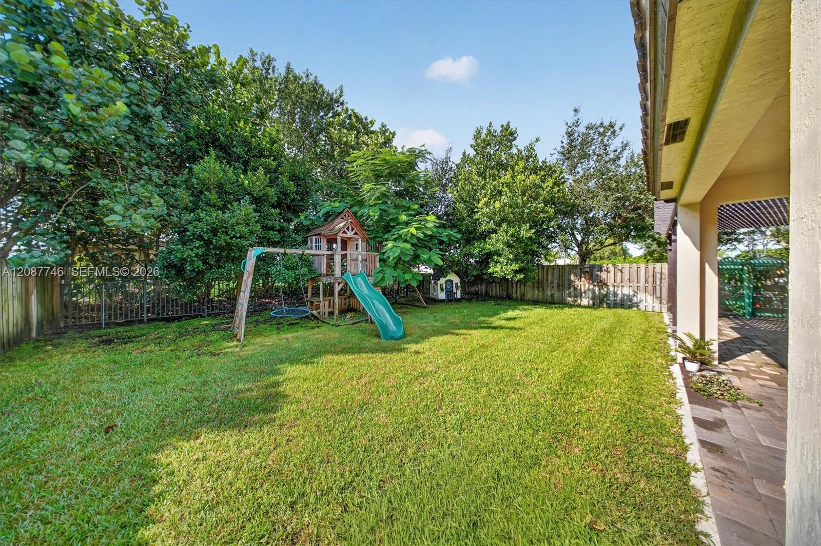 Backyard with a wooden playset and green slide, fenced lawn, mature trees, and paved patio.