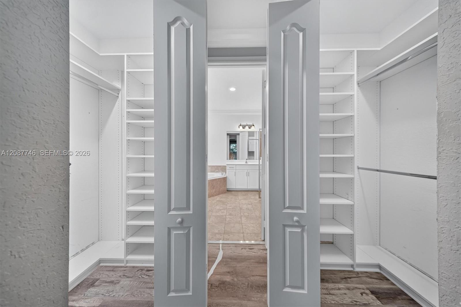 Walk-in closet with white built-in shelving, viewed through open gray doors toward a bathroom.