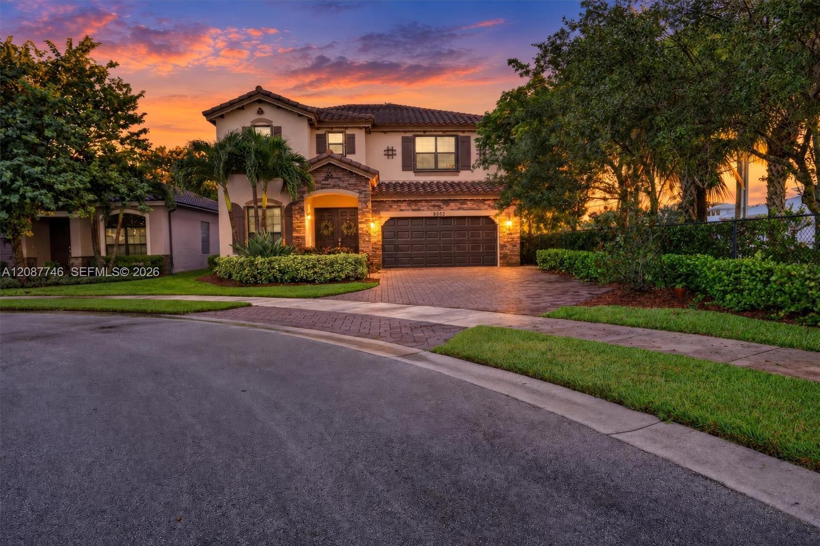 Two-story stucco home with a tiled roof and brick driveway, framed by palms at sunset.