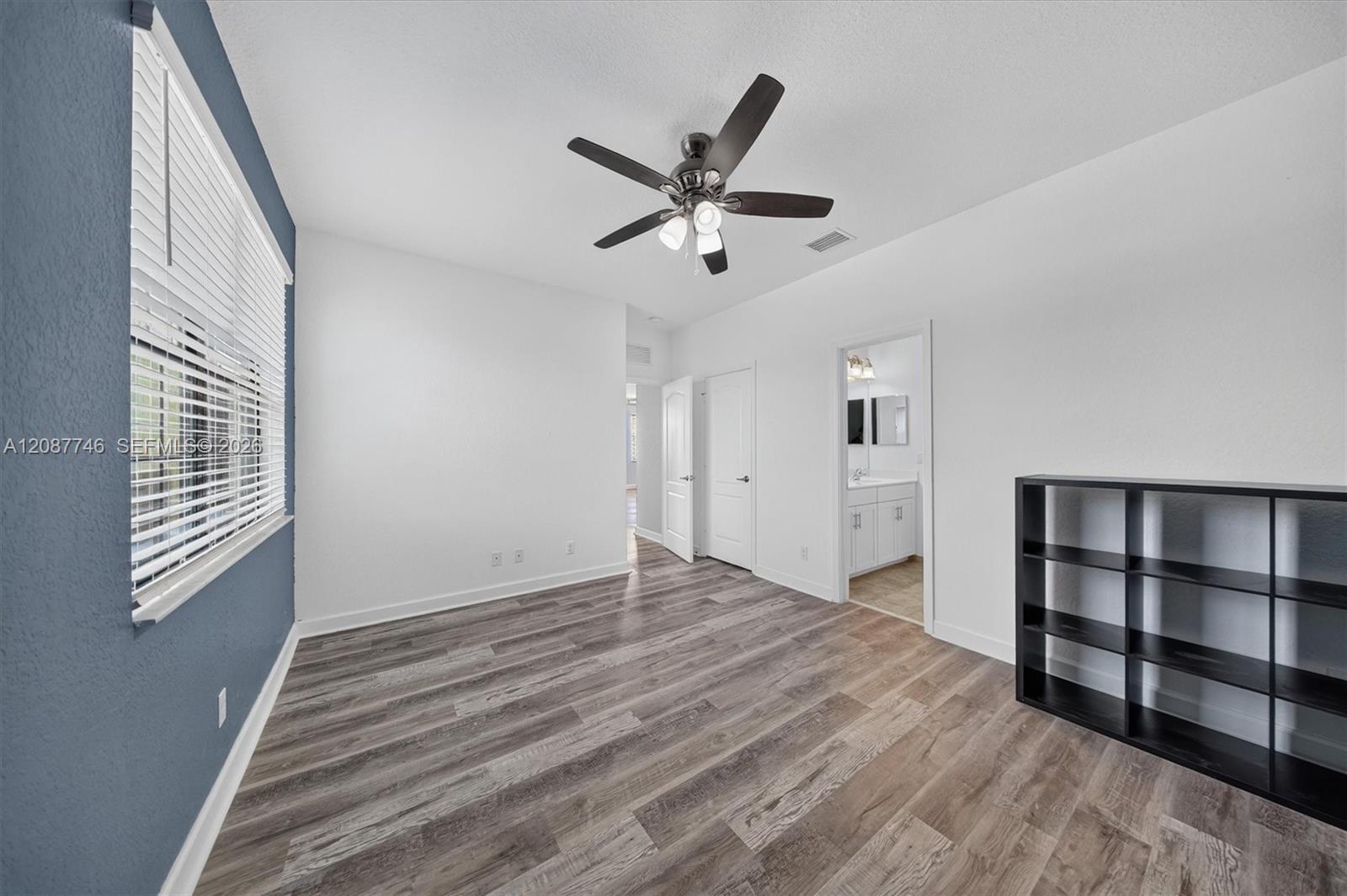 Empty bedroom with wood-look flooring, blue accent wall, ceiling fan, and doorway to a bathroom.