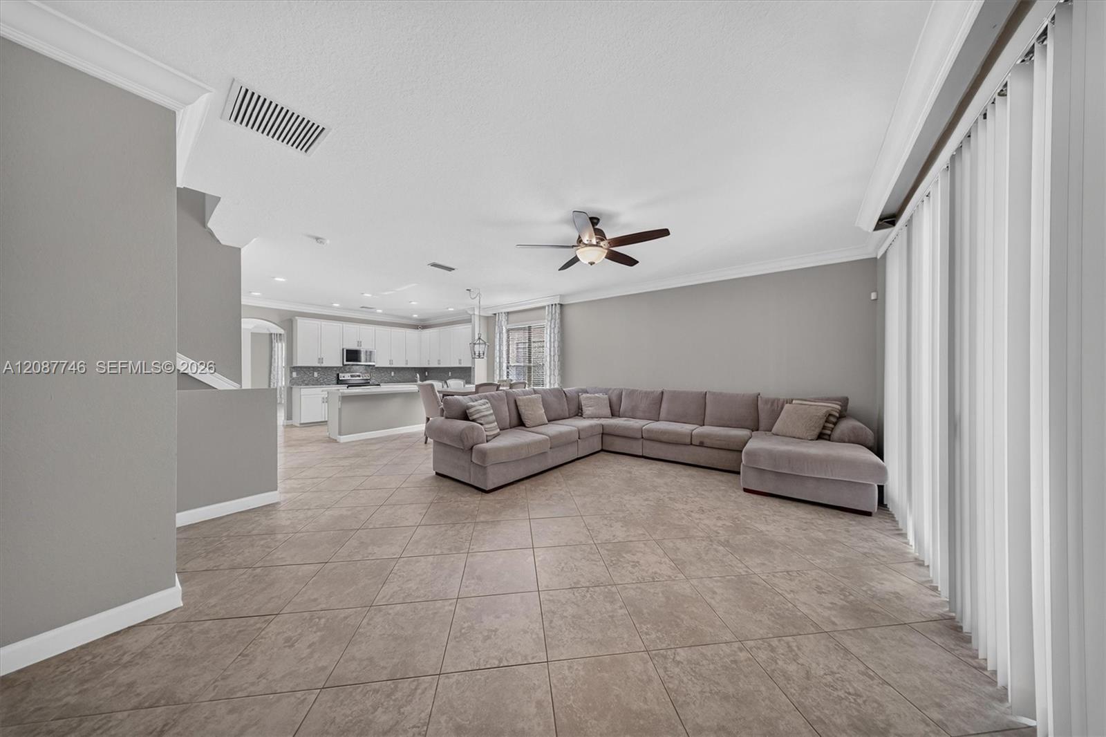 Living room with gray sectional sofa, large tiled floor, ceiling fan, and open kitchen.