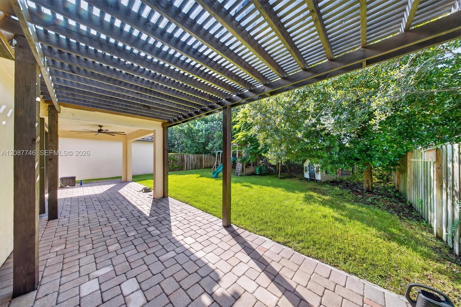 Backyard patio beneath a wood pergola, with a paver floor, fenced lawn, and children’s play equipment.