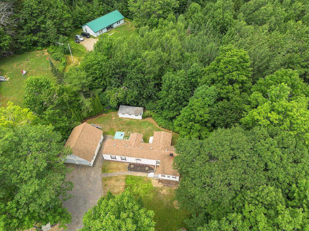 Aerial view of a home surrounded by dense woods, with a long driveway and a small outbuilding.