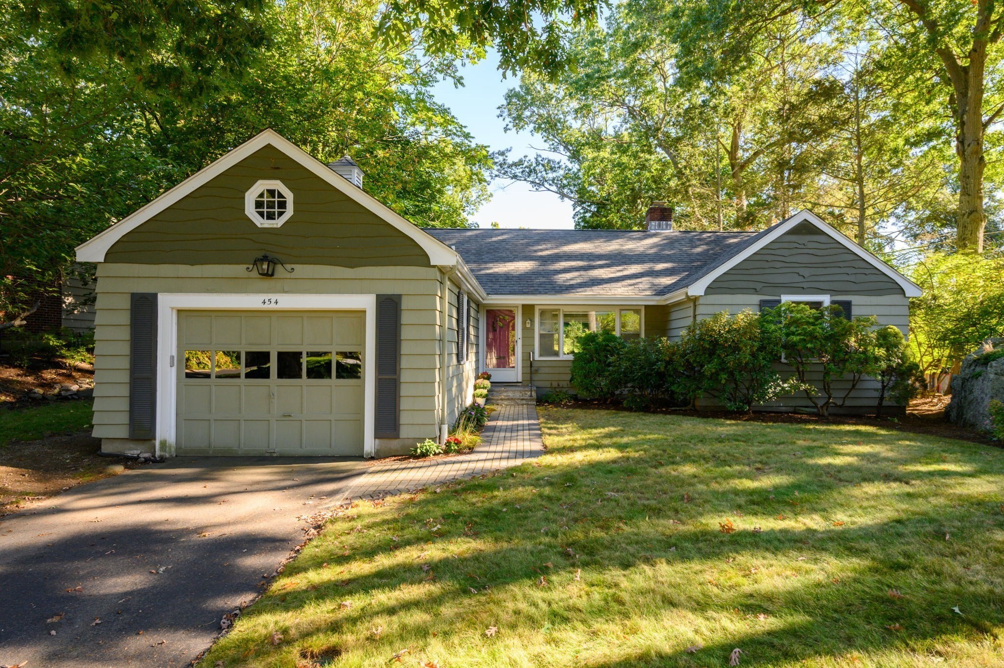 Gray-green single-story house with attached garage, red front door, and a brick walkway through a leafy yard.