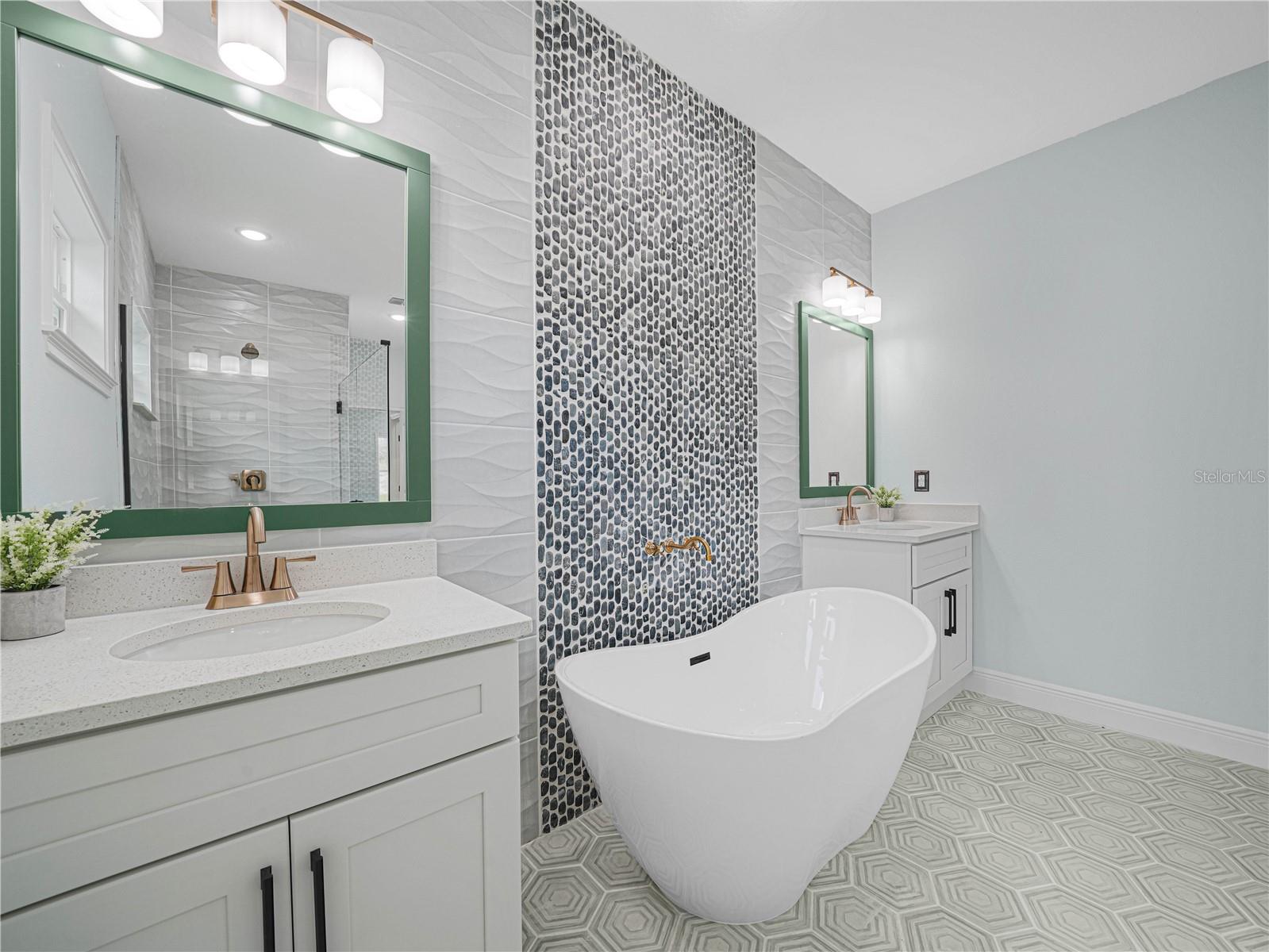 Modern bathroom with a freestanding soaking tub, pebble tile accent wall, and geometric patterned flooring.
