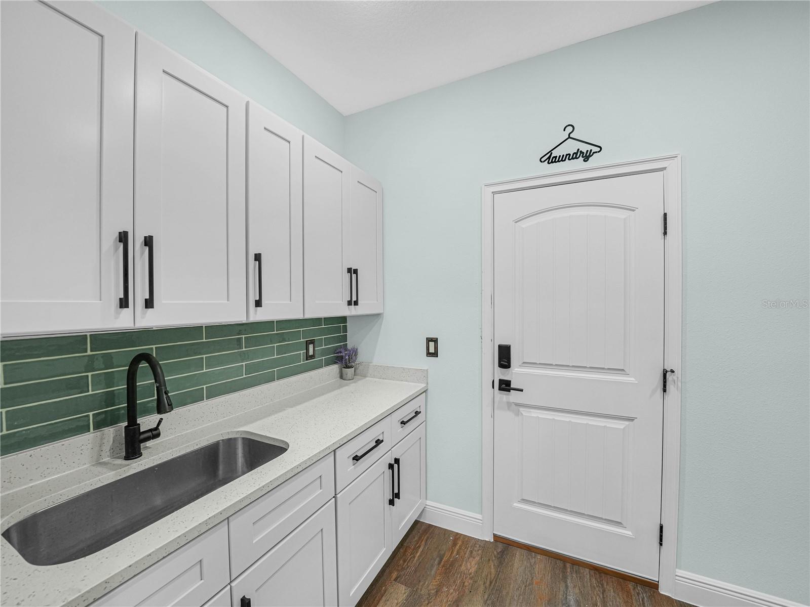 Laundry room with white cabinetry, green tile backsplash, and a deep stainless-steel sink.