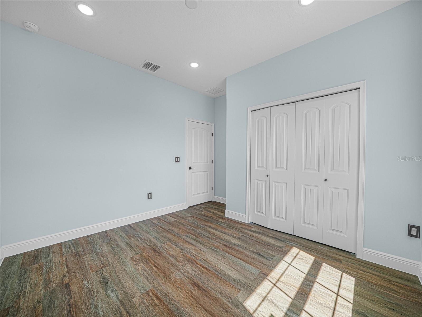 Empty bedroom with pale blue walls, white closet doors, and wood-look flooring.