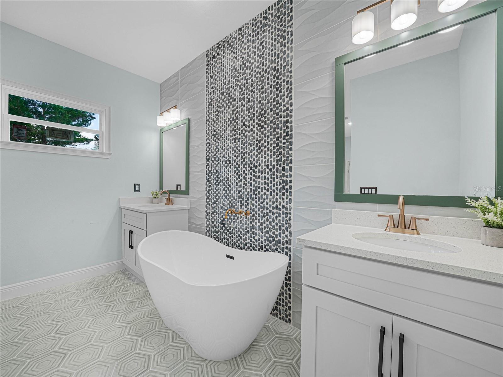 Bathroom with a freestanding tub, green-framed mirrors, pebble-patterned accent wall, and geometric tile floor.