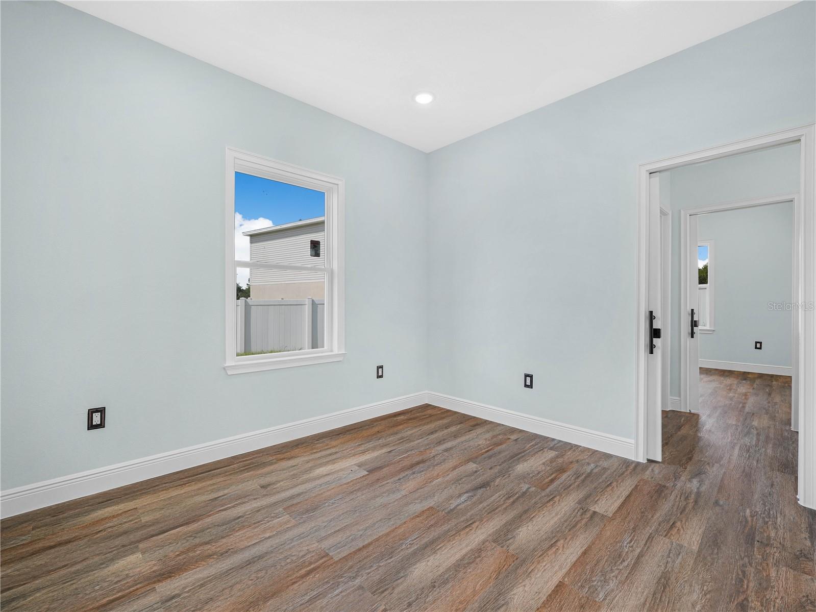 Empty room with pale blue walls, wood-look plank flooring, and a window overlooking a fenced yard.