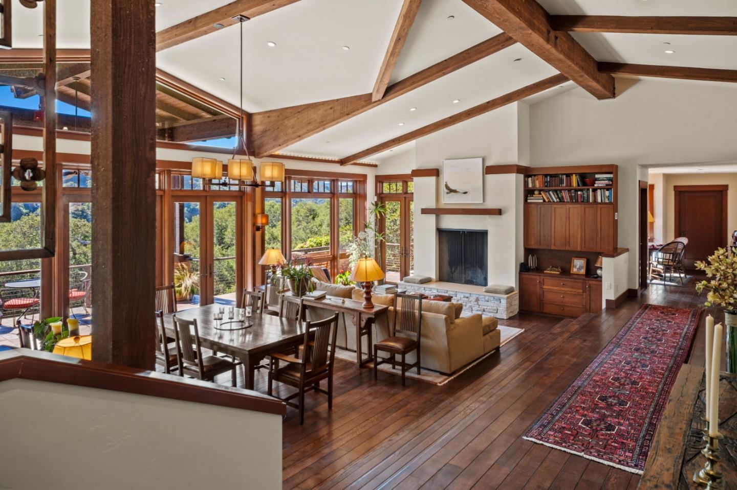 Open-plan living and dining room with exposed timber beams, a stone fireplace, and windows overlooking trees.